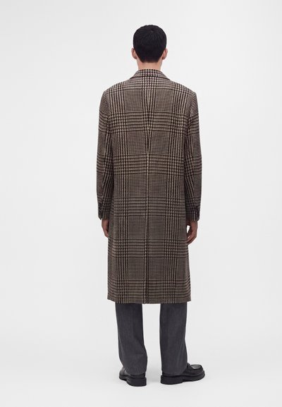 Homme de dos portant un long manteau à carreaux marron et noir, un pantalon gris et des chaussures noires sur fond blanc.