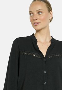 Woman with blonde hair in a bun wearing a black blouse with lace detail and buttons, looking to the side against a plain background.