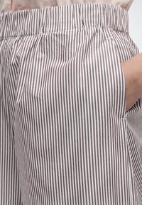 Main dans la poche d'un pantalon rayé avec des fines rayures verticales marron et blanches, taille élastique, et haut rose clair partiellement visible.