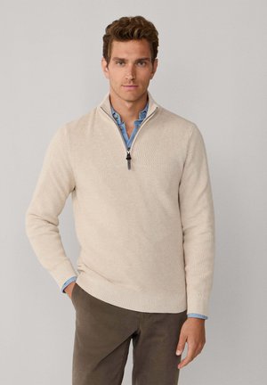 Suéter beige de canalé con cuello de media cremallera, presenta un diseño en forma de V y una textura suave. Usado sobre una camisa azul clara y combinado con pantalones oscuros.