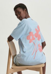 Lichtblauw katoenen T-shirt met een groot roze bloemenpatroon op de achterkant, korte mouwen en een relaxte pasvorm. Zittend op een houten stoel.