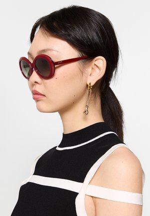 Vista laterale di una donna con capelli scuri raccolti in una coda di cavallo, che indossa occhiali da sole rotondi rossi, orecchini a catena d'oro e un top nero con strisce bianche.