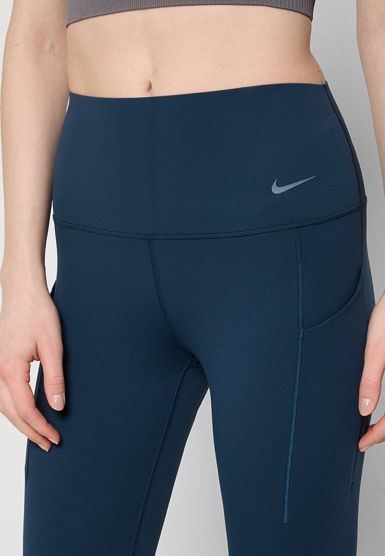 Mujer vistiendo leggings de Nike de tiro alto en azul marino con bolsillos laterales y una camiseta corta gris, mostrando la parte media del cuerpo y las manos.