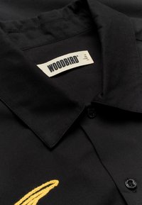 Chemise noire à col avec étiquette de la marque WOODBIRD cousue à l'intérieur du col et un petit motif jaune brodé près des boutons.