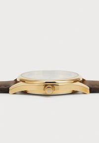 Montre-bracelet dorée avec un boîtier rond, un cadran en verre transparent et un bracelet en cuir marron. La couronne présente une accentuation de couleur claire.