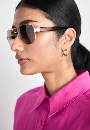 Brune tinnede solbriller med en guld metalramme og mønstrede brune tempelaccenter, båret af en person med lige mørkt hår og en lyserød skjorte.