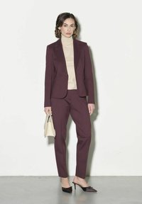Abito bordeaux con giacca aderente e pantaloni coordinati, abbinato a un dolcevita beige e mule nere con tacco, mentre tiene una borsa crema.