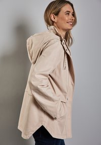 Veste de pluie imperméable beige clair avec capuche, fermetures à pression, poches latérales et un ourlet légèrement arrondi. La texture semble lisse, avec un drapé naturel.