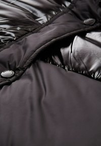 Zbliżenie na czarną kurtkę puchową, ukazującą matowe i błyszczące sekcje materiału z przeszywaną pikowaniem oraz dwoma widocznymi zapięciami marki.