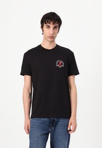 Black cotton t-shirt with a crew neck, featuring a small red and white logo on the chest, paired with blue denim jeans.