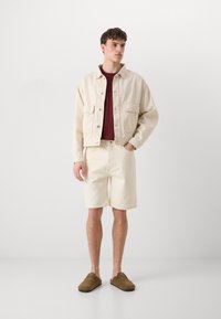 Vaalean beige farkkutakki napilla, yhdistettynä vaalean beigeihin shortseihin. Alle on puettu tummanpunainen T-paita. Ruskeat liukupohjakengät.