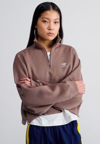 Sweatshirt castanho com fecho de correr até meio, de colarinho alto, com um pequeno logo branco e mangas oversized, usado sobre uma camisa branca.