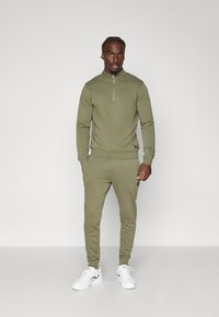 Olivengrønn treningsdress med glidelåskrage og ribbestrikkede mansjetter. Buksene har slim fit og lommer i siden. Båret med hvite sneakers.