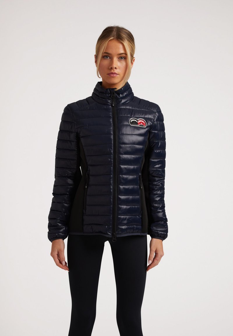 Marineblaue Steppjacke mit hohem Kragen, Reißverschluss und seitlichen Einsätzen; verfügt über ein Logo-Patch auf der Brust; glänzende Textur.