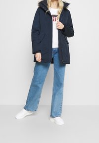 Tmavě modrá parka s odnímatelnou kožešinovou kapucí, zapínáním na zip, bočními kapsami a volným střihem, nošená přes džínové kalhoty a bílé tenisky.