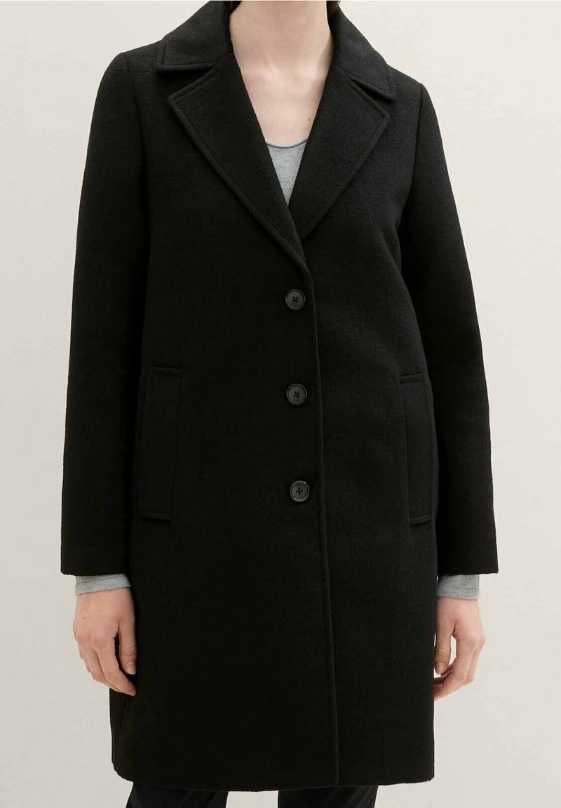 Personne portant un manteau en laine noir à trois boutons avec col à revers sur un haut à manches longues gris clair sur un fond uni.