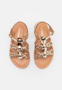 Sandales en cuir marron avec des brides métalliques tressées, boucle de cheville réglable et semelle intérieure lisse. Elles présentent un bout arrondi et un design minimaliste.