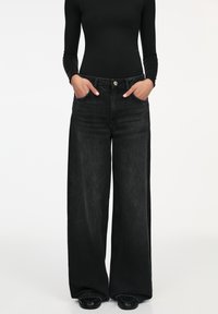 Jean denim noir à jambes larges avec une taille haute, des poches avant et une fermeture à bouton argenté. Présente une texture douce et un design classique.