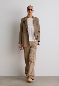 Béžový dvouřadý blazer, bílý top a světle béžové cargo kalhoty s bočními kapsami a volným střihem. Modelka nosí sluneční brýle.