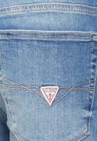 Jeans de denim azul claro con un bolsillo trasero que presenta un parche logo triangular rojo y blanco, costuras visibles y un acabado texturizado.