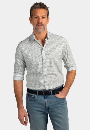 Man met kort haar en baard, gekleed in een wit overhemd met patroon en opgerolde mouwen en een blauwe spijkerbroek, die staat met één hand in zijn zak.