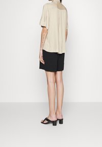 Personne debout de dos portant une chemise beige à manches courtes, un short noir et des sandales noires à talons sur un fond blanc.