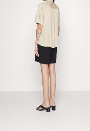 Osoba stojąca tyłem, ubrana w beżową koszulę z krótkim rękawem, czarne szorty i czarne sandały na obcasie na białym tle.