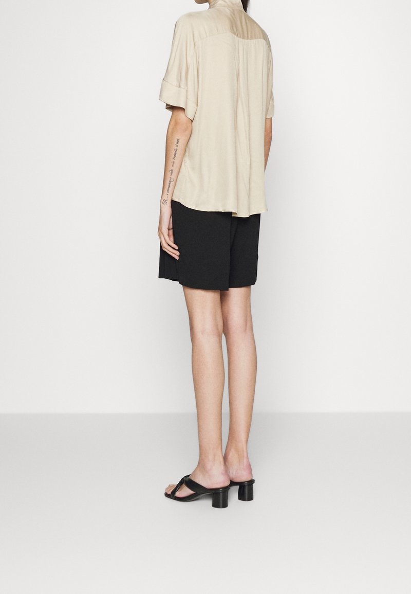 Personne debout de dos portant une chemise beige à manches courtes, un short noir et des sandales noires à talons sur un fond blanc.