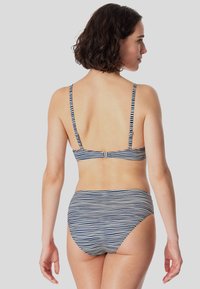 Pruhovaný bikini v námořnické modré a bílé barvě, s tenkými nastavitelnými ramínky a zapínáním na sponu na zadní straně. Materiál vypadá hladce a má přiléhavý design.