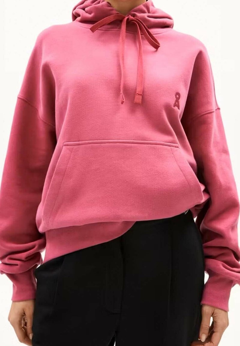 Personne portant un sweat à capuche rose ample avec poche avant et cordons, associé à un pantalon noir, montré du cou aux hanches.