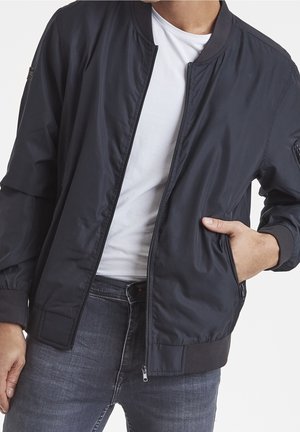 Man draagt een open zwarte bomberjack over een wit T-shirt met zijn hand in de jaszak, gecombineerd met donkerblauwe spijkerbroeken.