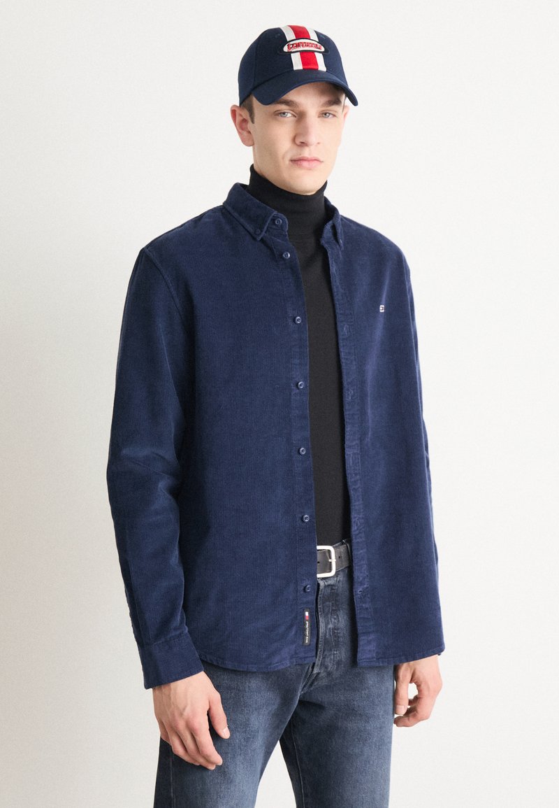 Camicia di velluto a coste blu navy con bottoni, abbinata a un dolcevita nero, jeans blu e un berretto blu con una striscia centrale e dettagli del logo.