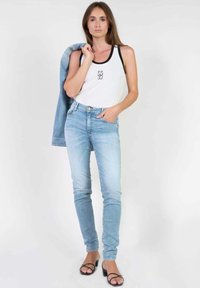 Witte tanktop met zwarte banden en logo, gecombineerd met lichtblauwe skinny jeans. Een jeansjack hangt over één schouder. Zwarte sandalen.