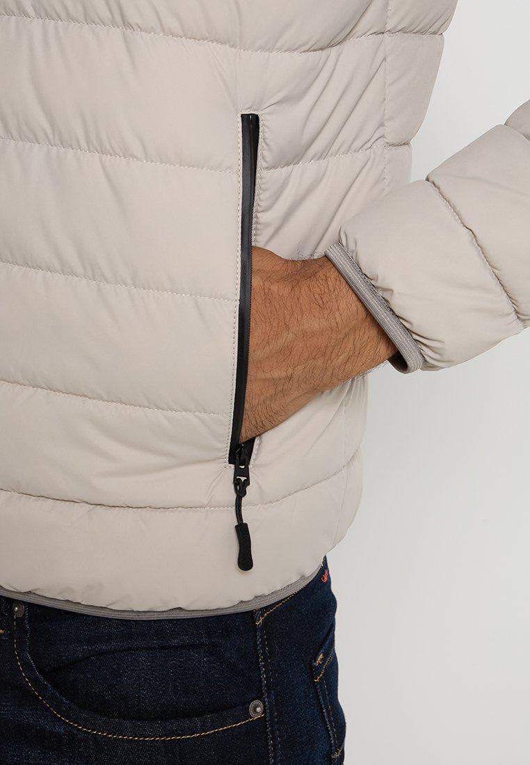 Hellgraue gepolsterte Jacke mit gestepptem Design, schwarzem Reißverschluss und hellgrauen Bündchen, getragen von einer Person mit der Hand in der Tasche.