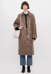 Long beige coat with a spotted pattern, large collar, and front buttons. Paired with dark top, blue jeans, and black shoes.  Holding a white bag.