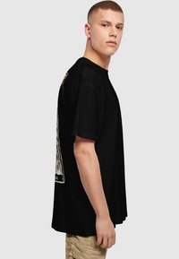 Sort bomulds t-shirt med korte ærmer, der har et stort grafisk print på ryggen. Løs pasform, rund halsudskæring, glat tekstur.