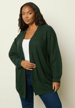 Vrouw met een donkergroene gebreide cardigan over een witte top en blauwe jeans, poseert met één hand op de cardigan voor een neutrale achtergrond.
