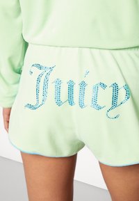 Světle zelené šortky s modrým kamínkovým nápisem "Juicy" na zadní straně, které má na sobě osoba s opálenou pletí.