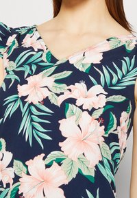 Námořnický květinový top s růžovými květinami hibiscus a zelenými listy, s výstřihem do V a zlatým lemem kolem výstřihu. Měkká textura látky.