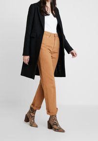 Žena v černém dlouhém kabátě, bílém topu, vyhrnutých krémových kalhotách a kotníkových botách s hadím vzorem na podpatku kráčí na jednobarevném pozadí.