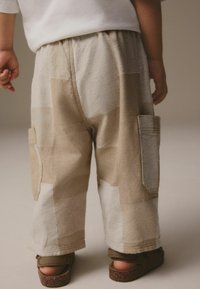 Beige Patchwork-Hose mit lockerem Schnitt, elastischem Bund und zwei Seitentaschen. Texturiert mit kontrastierenden hellen und dunklen Patches.