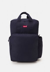 Tamsiai mėlynas audinio kuprinė su dviem rankenomis, priekiu kišenė ir šonine tinkline kišene. Priekyje yra raudonas Levi's logotipas.