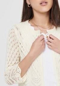Light cream knitted cardigan featuring an open weave pattern and three-quarter sleeves, worn over a white top, with a round neckline.