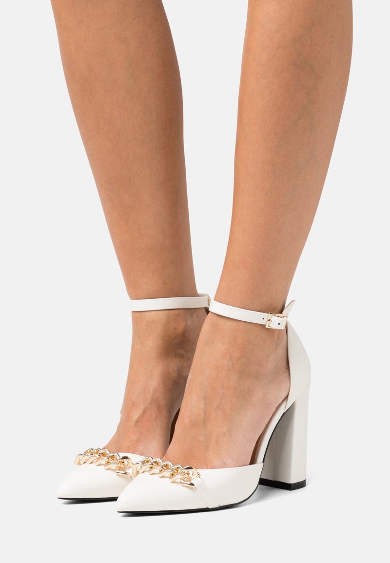Chaussures blanches à talons hauts avec brides à la cheville et détail chaîne dorée portées pieds nus sur un fond uni.