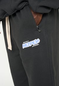 Zwarte joggingsbroek met een geborstelde textuur, voorzien van een zijritszak en een wit en blauwe logo met de tekst "MULTIPLY APPAREL."