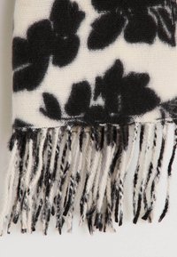 Patrón floral negro sobre una bufanda crema, hecha de material suave, con bordes con flecos en tonos negro, blanco y gris.