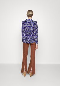 Blouse fleurie aux tons bleus avec manches bouffantes, associée à un pantalon évasé marron avec fentes latérales, complété par des talons à brides.