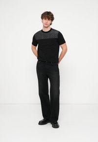 T-shirt preta com painel texturizado preto no peito, combinada com jeans escuros de perna reta e sapatos pretos. Design simples e casual.