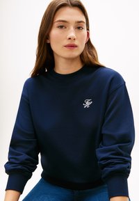 Μπλε σκούρο φούτερ με ριμπ μανσέτες και τελείωμα, με μικρό λευκό κεντημένο λογότυπο στο στήθος, κατασκευασμένο από μαλακό, υφασμάτινο ύφασμα με υφή.