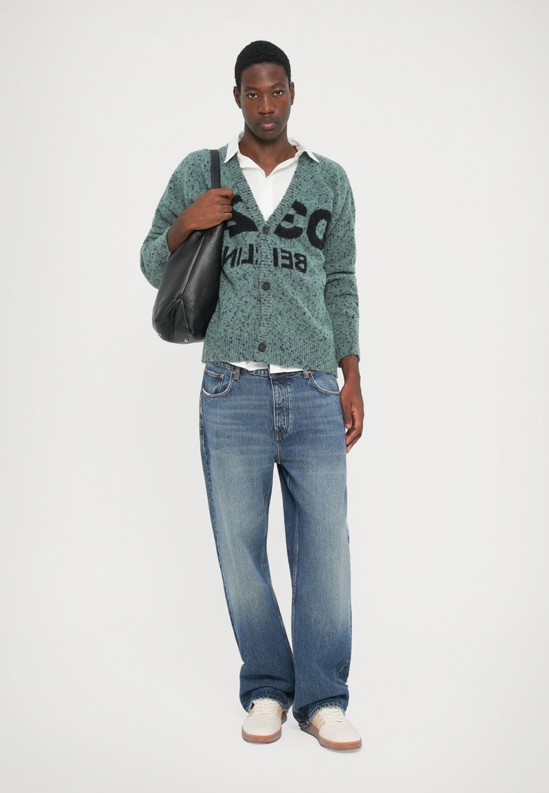 Green speckled cardigan with black logo, white collared shirt, loose blue jeans, and white shoes. Black shoulder bag accentuates outfit.